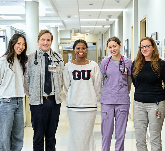 A group of five people stand together in a brightly lit hospital hallway, smiling at the camera. Two wear stethoscopes and medical attire, one wears a Gonzaga University sweatshirt, and others are dressed in casual or professional clothing, representing a mix of students and healthcare professionals.