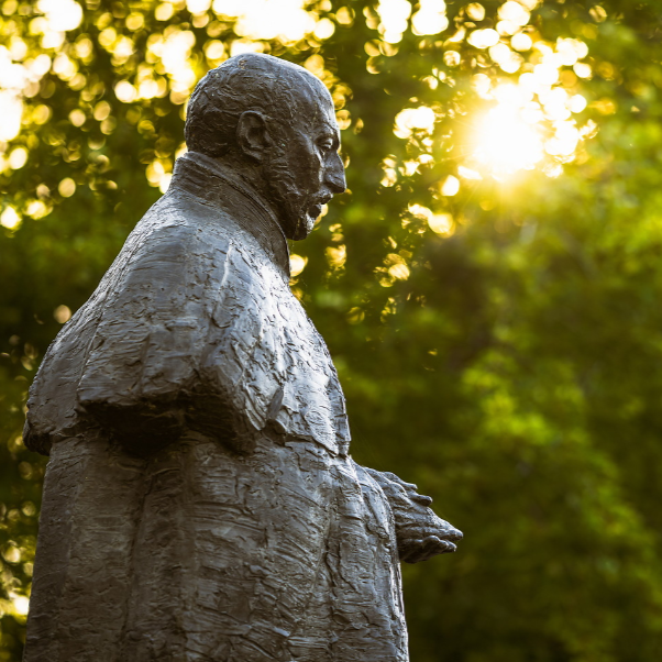 Profile view of a black statue of St. Ignatius of Loyola with sunshine streaming through the trees in the background in front of College Hall.