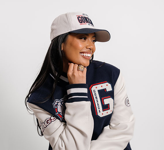 Keana Delos Santos in front of a white background wearing a Gonzaga coat and hat with her playoff ring on her hand.