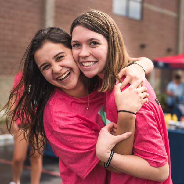 Two female students hugging each other. Both are wearing light red tee shirts are are smiling. The girl on the left has wavy black hair and the girl on the right has short light-brown hair. In the background is a brick building and other people.