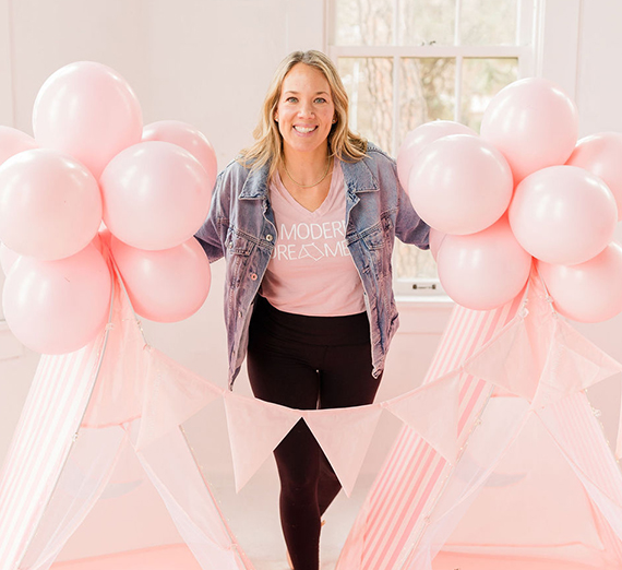 A woman stands between two pink tents with balloons.