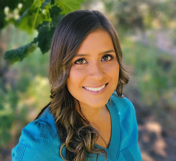 April O'Hair MBA alumna wearing blue v-neck, brown hair curled and down, with background of greenery