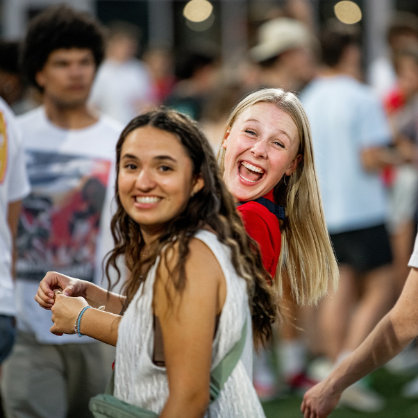 Two female students smiling at the camera hanging out with each other. The girl in the foreground has wavy dark brunette hair and is wearing a white tank top. The girl behind her has long straight blonde hair and is wearing a red tee shirt. People are walking around in the background.
