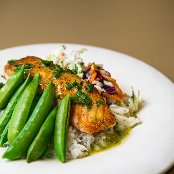 A picture of a plate of food at the COG on a tan table and white plate. The plate has the famous COG salmon on top of white rice and is accompanied by snap peas and spices on top of the salmon.