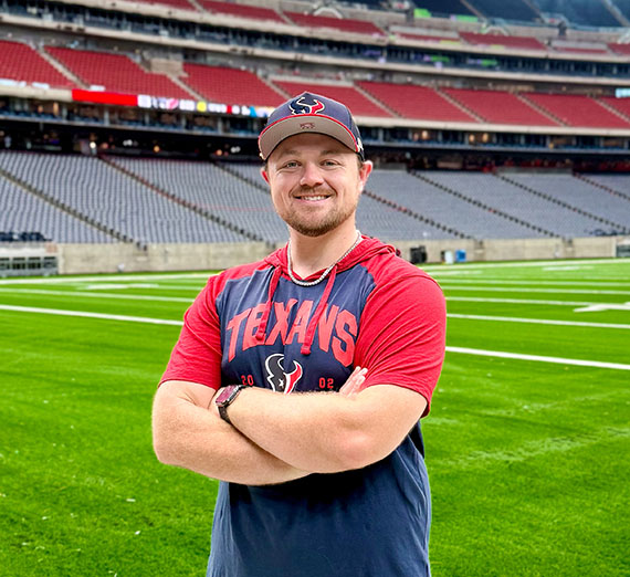 White man wearing baseball hat and Houston Texans tshirt standing with football field and stands in background