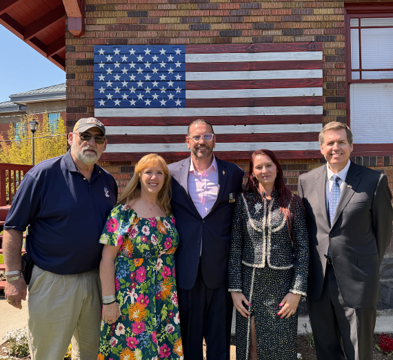 Five people stand in a row in front of  a flag.