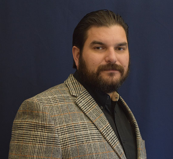 Brown haired man with beard, Shiloh Freijo, wearing blazer and black button up standing in front of blue curtain