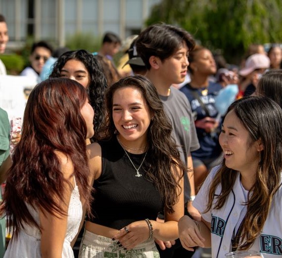 Three students smiling at each other while attending the Ӱԭ Club Fair. They are three female students who are laughing to each other with a crowd of people around them also at the club fair.