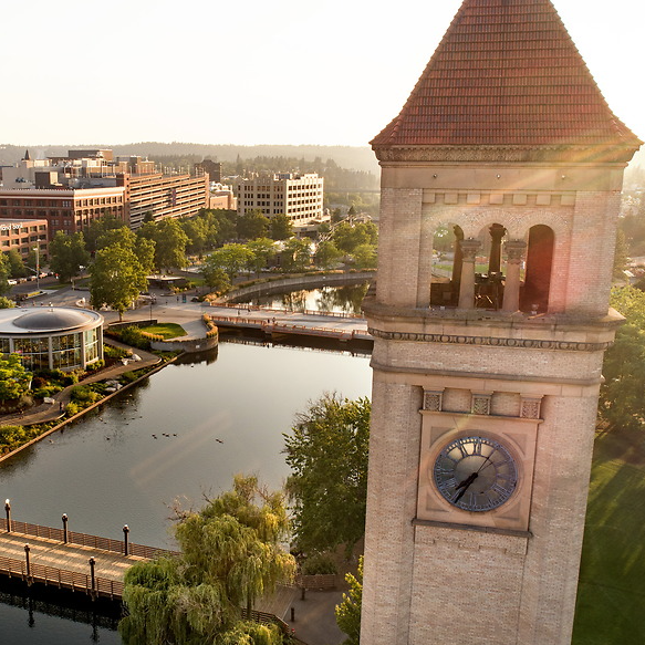 A photo of the view overlooking a clocktower and the Spokane River running through Downtown Spokane. The clocktower is on the right in the foreground and is made out of concrete and brick with a clock built into the side. There is a bridge over the river with people walking across in the lower left-hand side of the photo. In the background, there is a view of the downtown area of the city of Spokane. 