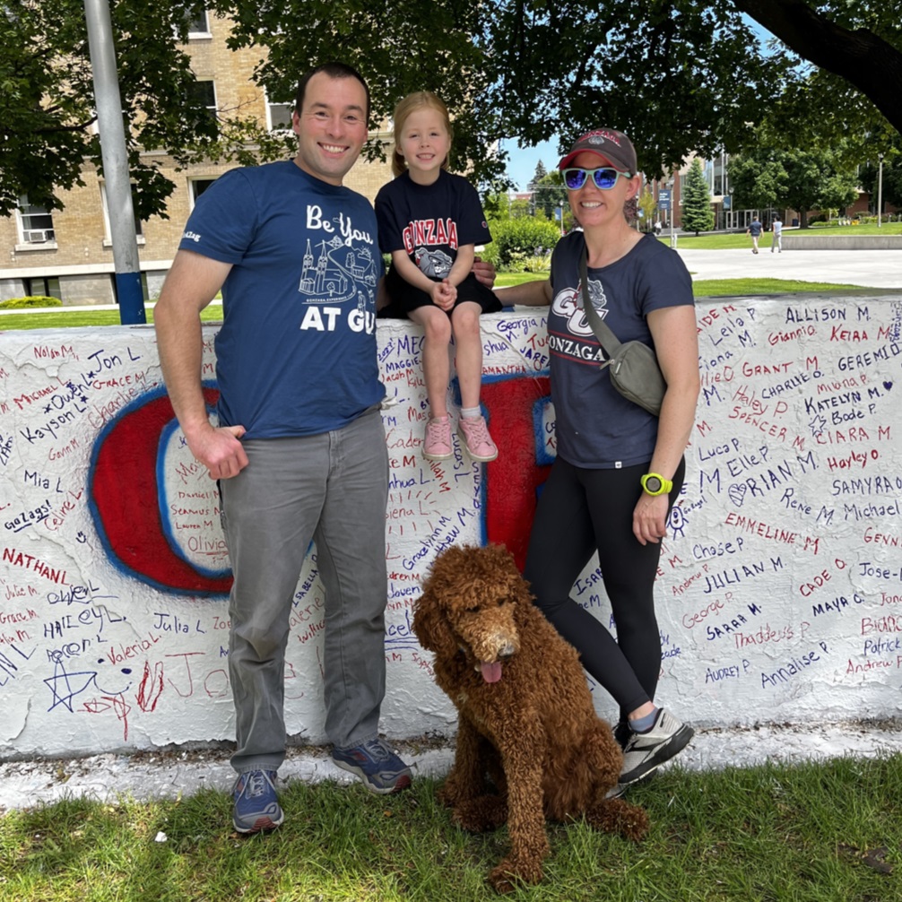 Stephen Keller, Senior Director of Undergraduate Admission, standing in front of signed white wall with his daughter sitting on top of the wall, wife standing next to him, and curly-haired dog sitting down beside him.