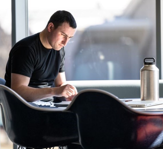Male student wearing a black t-shirt, sitting at a table in the Bollier Center writing in a notebook.