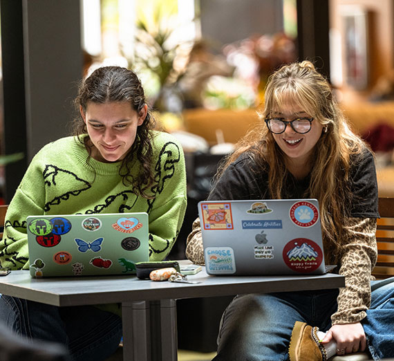 two female students sit at a table with laptops