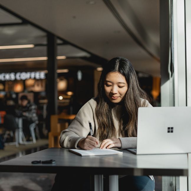 Student writing on a notebook in the Hemmingson Center. The female student is wearing a beige sweater and has long black error and is smiling as she writes. To her left is her silver laptop on top of a table.