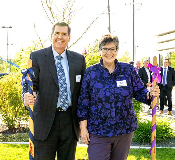 two individuals stand side by side with shovels, one wrapped in blue and the other in purple