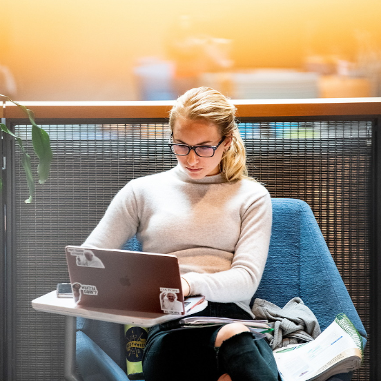 Student studying in Hemmingson Center who is a woman with blonde hair and a grey turtleneck sweater with black glasses. She is typing on her laptop and sitting in a blue armchair next to a plant.