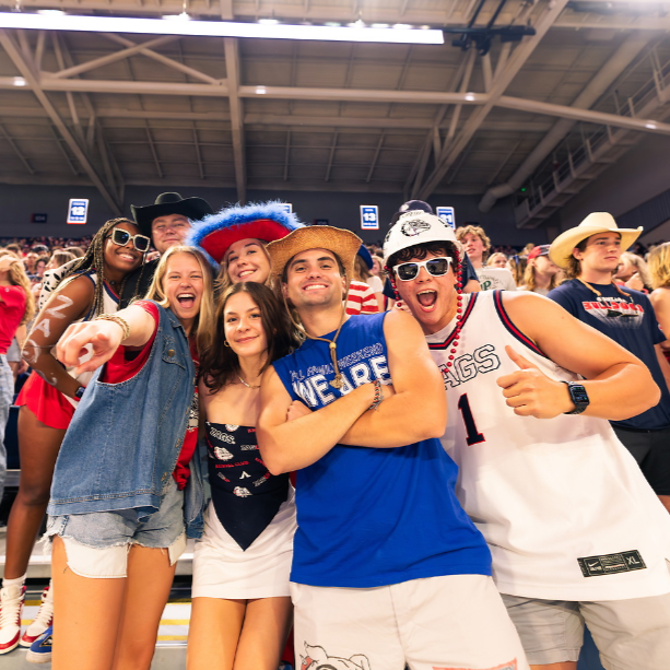 Gonzaga University standing in the Kennel's student section for a basketball game. Students are smiling at the camera and dressed in red, white, and blue clothing.