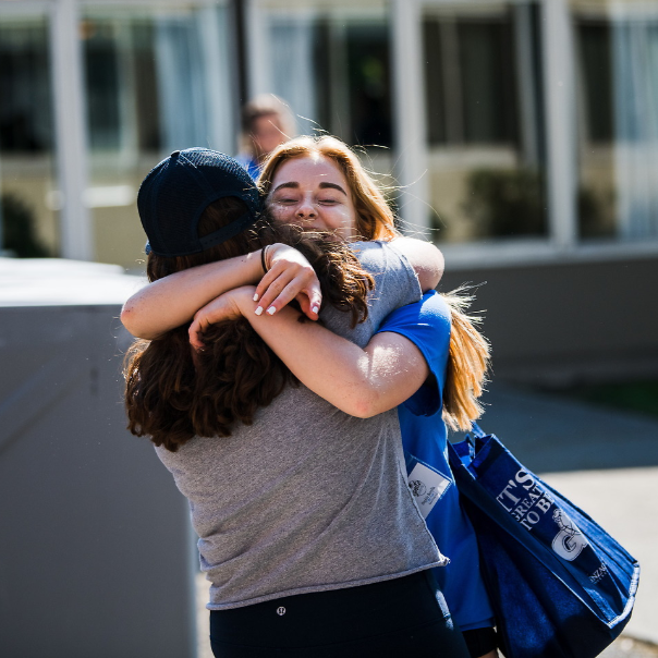 Two women hugging each other. The woman facing the camera has red hair and is smiling wearing a blue tee shirt. The woman with her back to the camera has dark wavy hair and is wearing a grey tee shirt and a navy hat.