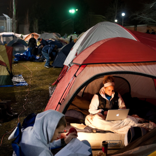 A girl in a white sweatshirt and a black puffer vest sits in a red tent in the foreground. She is working on a laptop and has a white blanket around her. In front of her, is a man in a blue lawn chair in a grey sweatshirt with the hood pulled up looking down at a piece of paper. In the background, there is a grass and many other tents behind the red tent. It is at night and the area is lit by streetlamps in the background.