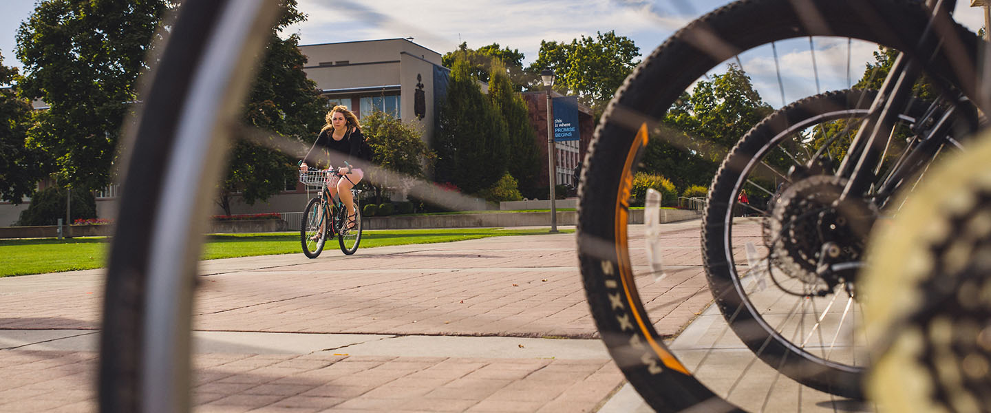 Student biking on campus, viewed through the spokes of a bike wheel.