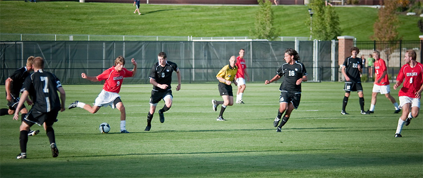 The image shows a soccer match being played outdoors on a grass field. Players are wearing uniforms: one team is in red jerseys (with the word "GONZAGA" visible on one), and the other team is in black. A referee in a yellow shirt is also present. The player in red (#9) appears to be preparing to kick the ball, while multiple players from the opposing team are moving toward him to defend. Spectators or staff can be seen in the background.