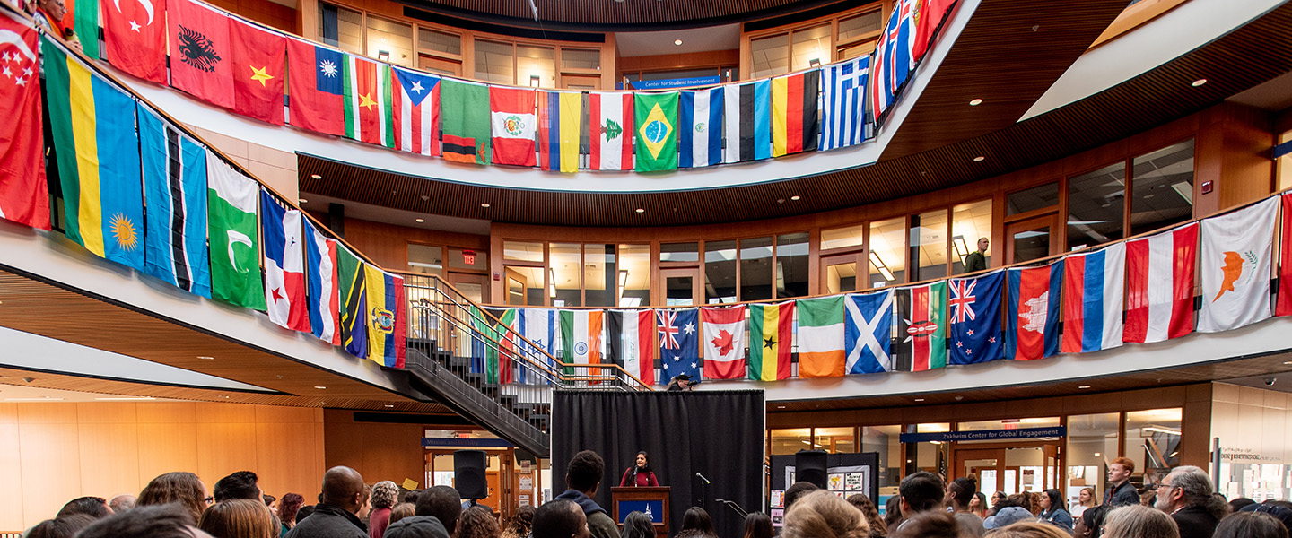 Decorative image, group gathered to hear a speaker. Global country flags hang off balcony.